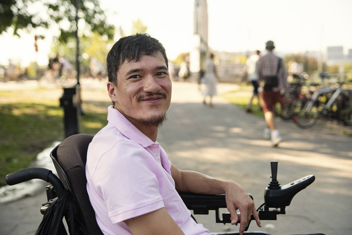 Portrait of asian man living with cerebral palsy, outdoors in summer. He is wearing a pink polo, sitting on a wheelchair that is on a sidewalk. Vertical waist up outdoors shot with copy space.