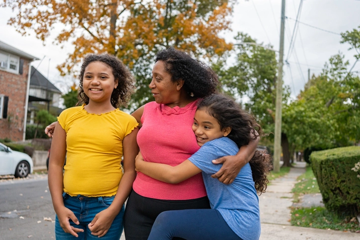 A woman stands on a neighborhood sidewalk with two young girls, smiling and embracing each other.