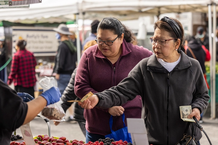Two women shop for fresh berries at an outdoor farmers market, purchasing produce from a vendor at a community market stand.