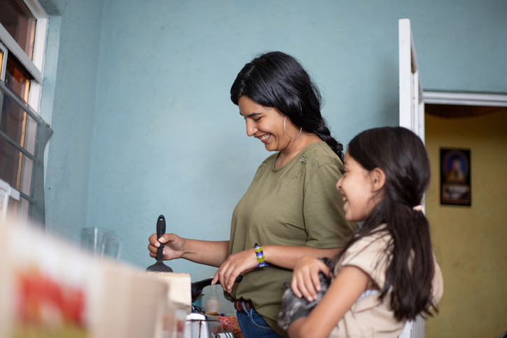Mexican thirtysomething woman cooking in her modest kitchen, with her daughter