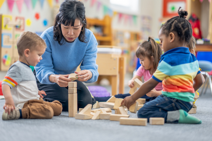 A preschool teacher sits on the floor with a small group of students as they build together with wooden blocks. They are each dressed casually and are focused on the activity.