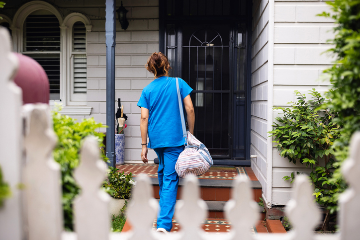 A female nurse in blue scrubs is walking towards a house while carrying a medical bag. The scene depicts a professional role, commitment to health care, and a caring attitude, possibly during a home visit.