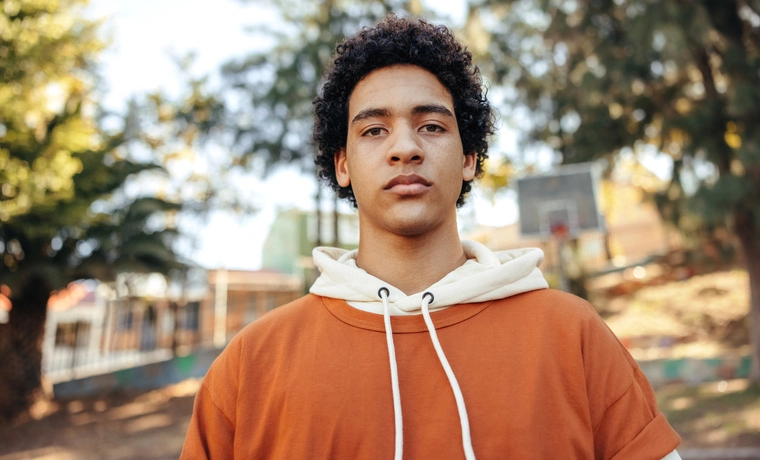 Teenage boy looking at the camera outdoors during the day. Fashionable teenager wearing casual clothing in an urban park. Confident male youngster standing alone outdoors.