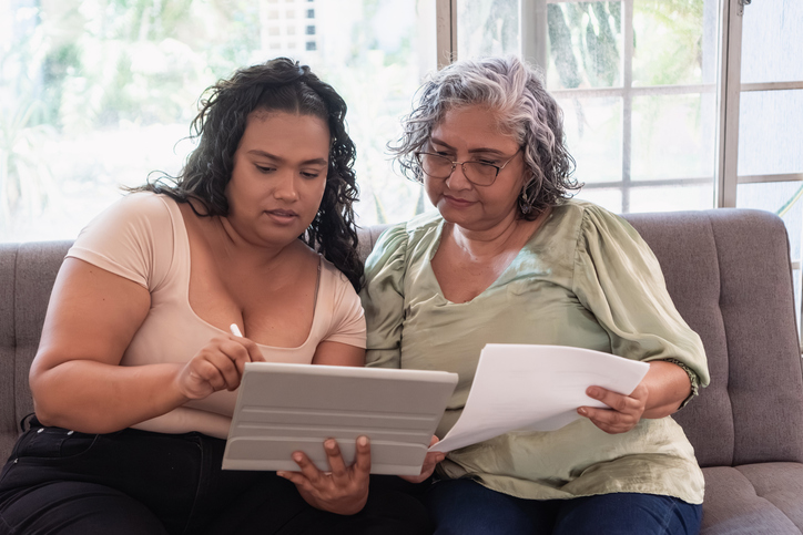 Young adult woman helping senior woman understand documents and technologies at home