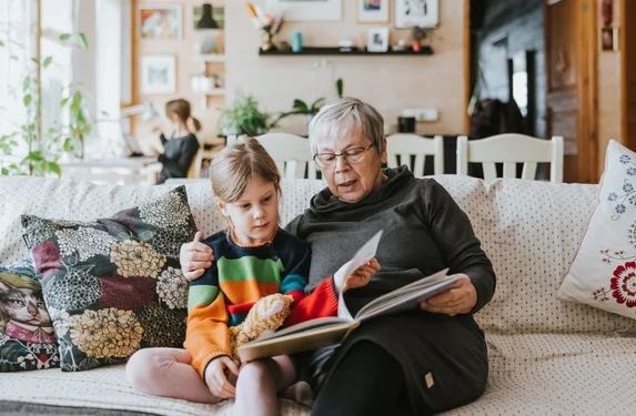 An older adult and a young child sit together on a couch in a cozy living room, looking through a photo album or book. The older adult points to a page while the child follows along, suggesting a moment of shared learning, storytelling, or family connection. A second person is visible in the background near a window.