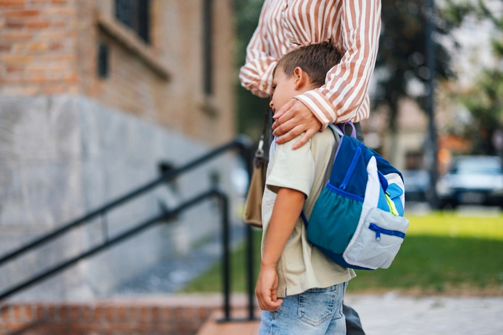 A young child wearing a blue backpack hugs and adult's waist outside a school building, capturing a quiet moment during school drop-off.
