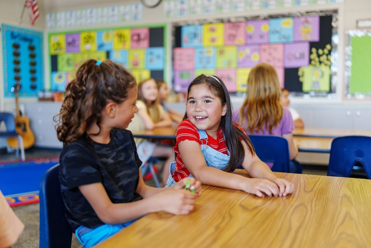 A cute Eurasian girl sits with a friend at a desk in their classroom and talks cheerfully during class.
