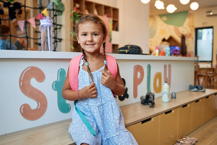 Little girl at the kindergarten. She's sitting down, wearing a blue dress and has a backpack on her shoulders. Back to school concept.