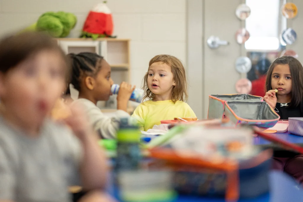 Diverse preschool children sit at a colorful table eating lunch and drinking from bottles in a bright classroom, showing friendship, routine, and early childhood learning in a school setting