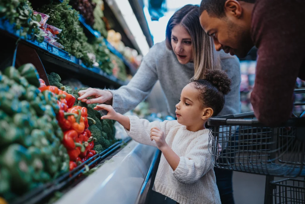 A biracial couple picks out vegetables with their preschool daughter's help while shopping at a grocery store.